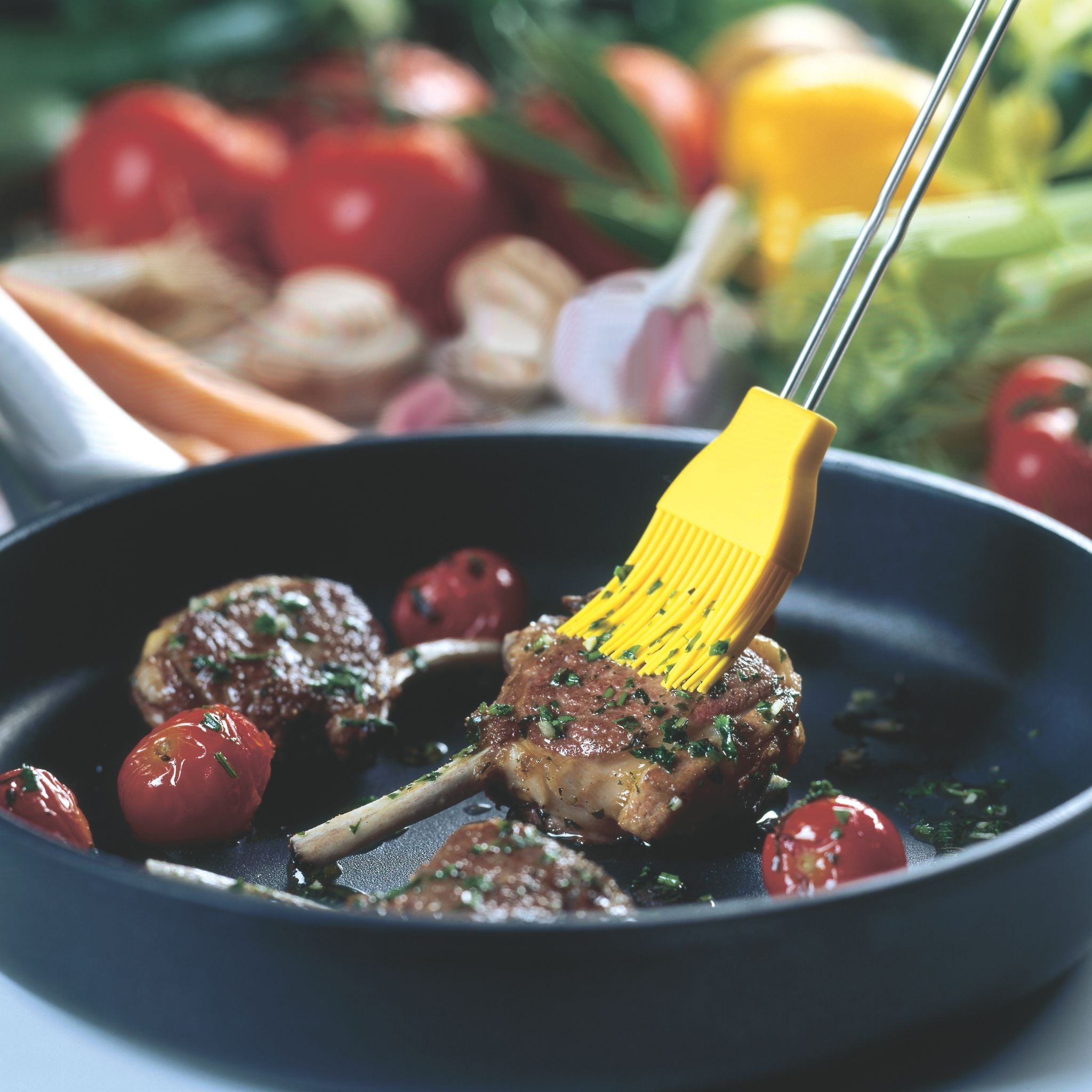 Schwarze Pfanne mit gebratenem Lammkotelett, Kirschtomaten und gelbem Silikonpinsel.