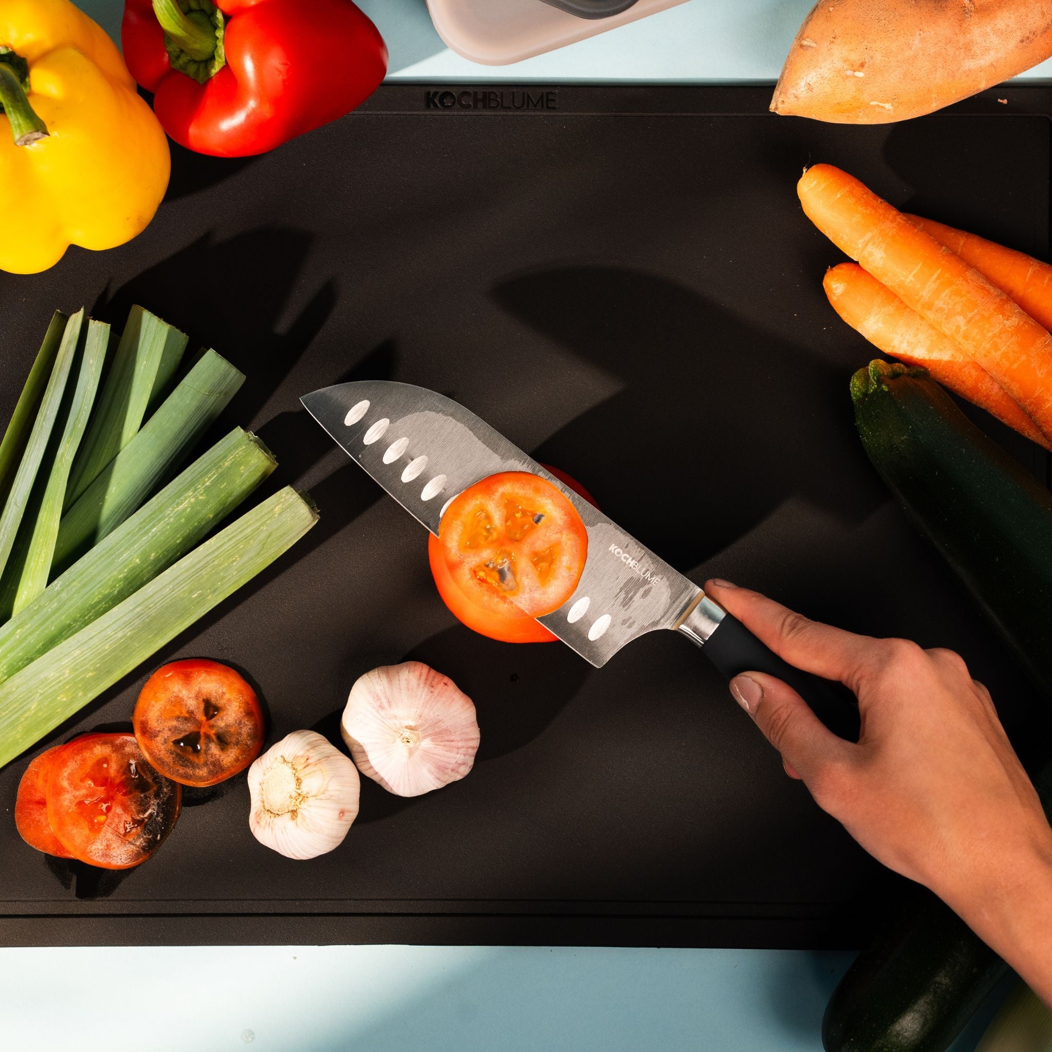 Schwarze Schneidunterlage mit Gemüse und einem Messer, das eine Tomate schneidet.