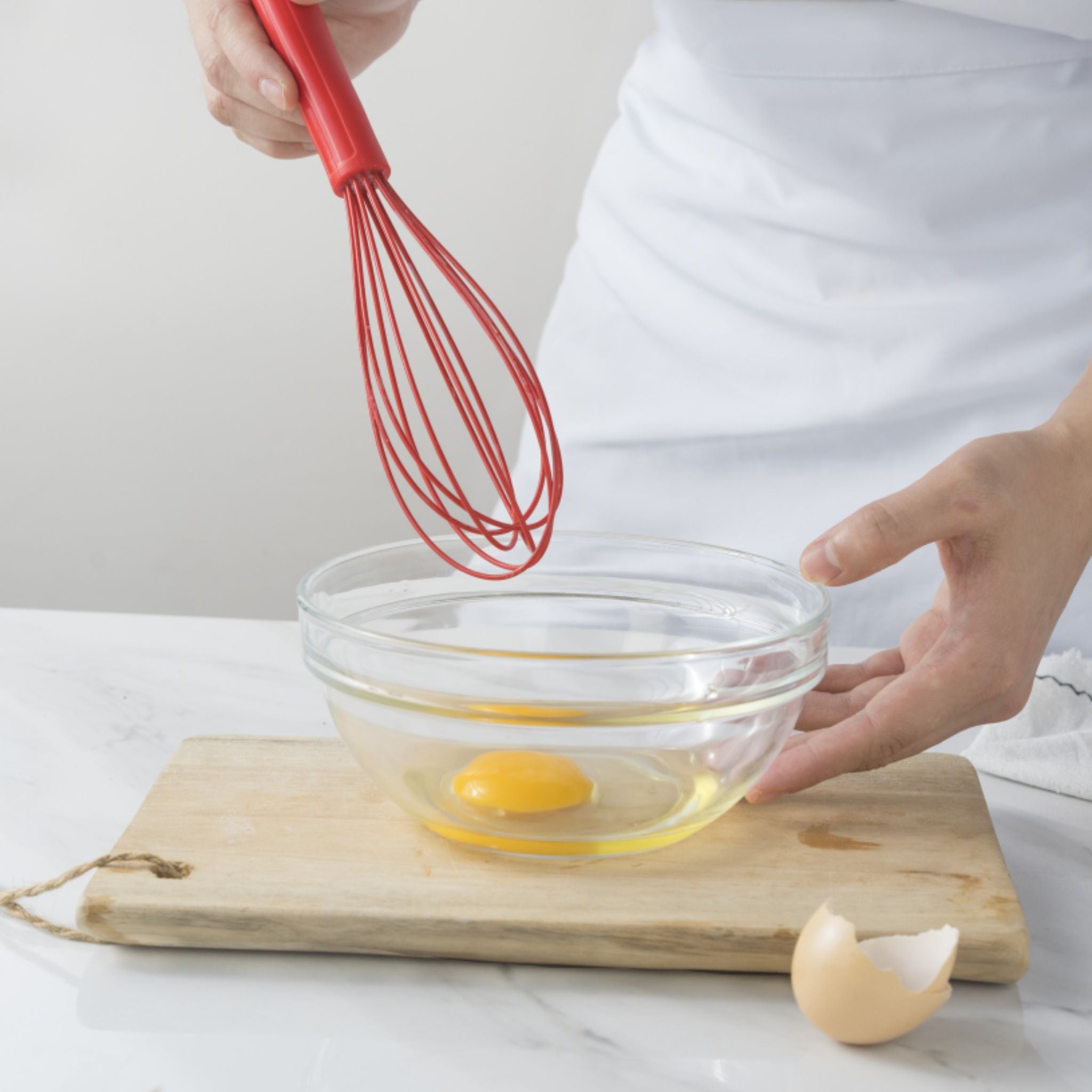 Roter Schneebesen aus Silikon schlägt Ei in Glasrührschüssel auf Holzbrett.