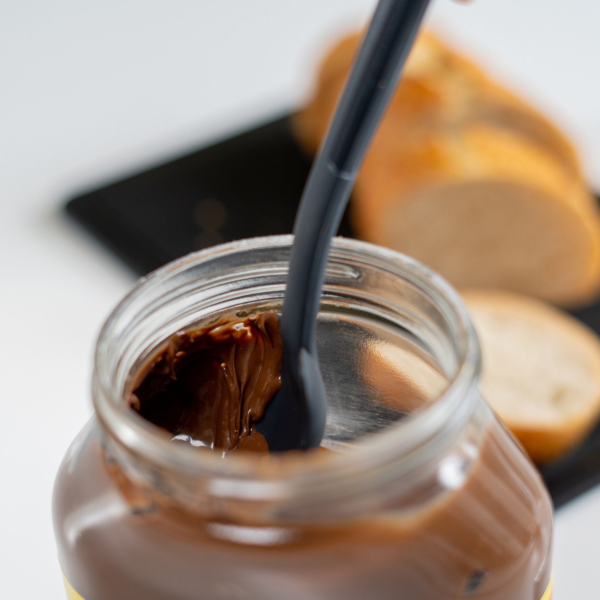 Anthrazitfarbener Restelöffel rührt in offenem Glas mit Schokoladecreme, Baguette im Hintergrund.