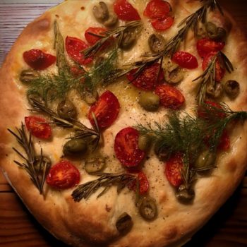 Ein fertiges Focaccia mit Tomaten, Rosmarien und Oliven auf einem Hozbrett.