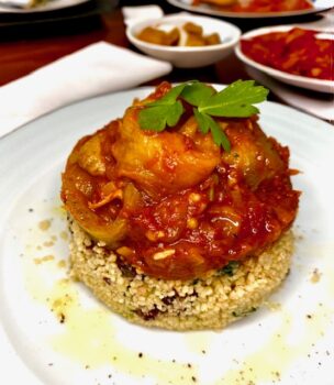 Auberginenauflauf mit Couscous in Servierring auf weißem Teller angerichtet und mit etwas OIivenöl und Pfeffer dekorativ verfeinert.