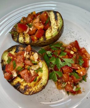 Zwei gegrillte Avocado mit Tomaten-Salsa gefüllt auf einem weißen Teller mit etwas Tomaten-Salsa und Petersilie angerichtet.