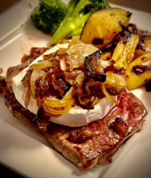 Ein Camembert-Steak mit einem ganzen runden Camembert auf dem Steak mit gerösteten Zwiebeln. Im HIntergrund zwei Röschen vom Brokkoli und angebratene Kartoffelspalten auf einem Teller angerichtet.