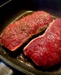 Zwei Stück Fleisch in einer Pfanne. Das eine Stück Fleisch ist gewürzt mit Salz und Pfeffer - das andere Fleisch ist ungewürzt.