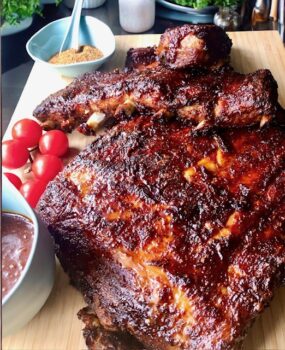 Spareribs auf einem Holzbrett mit Tomaten und BBQ-Sauce angerichtet.