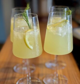 Limoncello Spritz in vier Gläsern angerichtet und mit Zitrone und einem Zweig Rosmarien garniert.