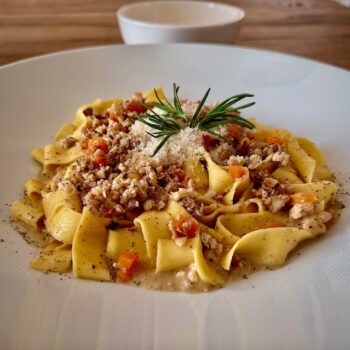 Breite Nudeln mit einem hellen Ragù auf weißem Teller mit Parmesan und Rosmarin dekoriert.