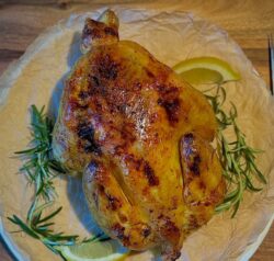 Ein ganzes leicht gebräuntes Huhn aus dem Ofen auf einem Teller mit Rosmarien und Zitrone angerichtet.