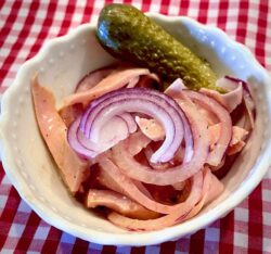 Leberkäs-Wurstsalt in einer kleinen weißen Schüssel mit einer sauren Gurke und roten Zwiebeln angerichtet.