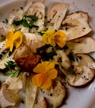 Auf einem Tisch liegt ein weißer Teller mit Steinpilz Carpaccio, dekoriert mit essbaren Blumen und Parmesan.