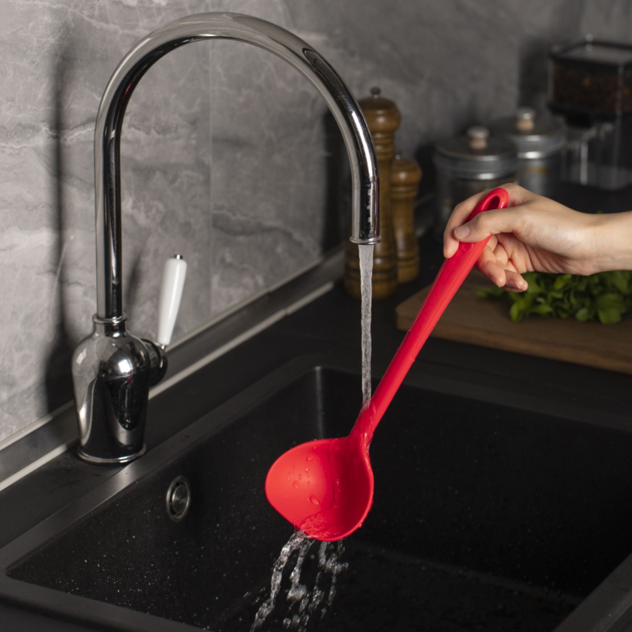 Rote Schöpfkelle aus Kunststoff mit langem Stiel über schwarzer Spüle unter laufendem Wasserhahn.