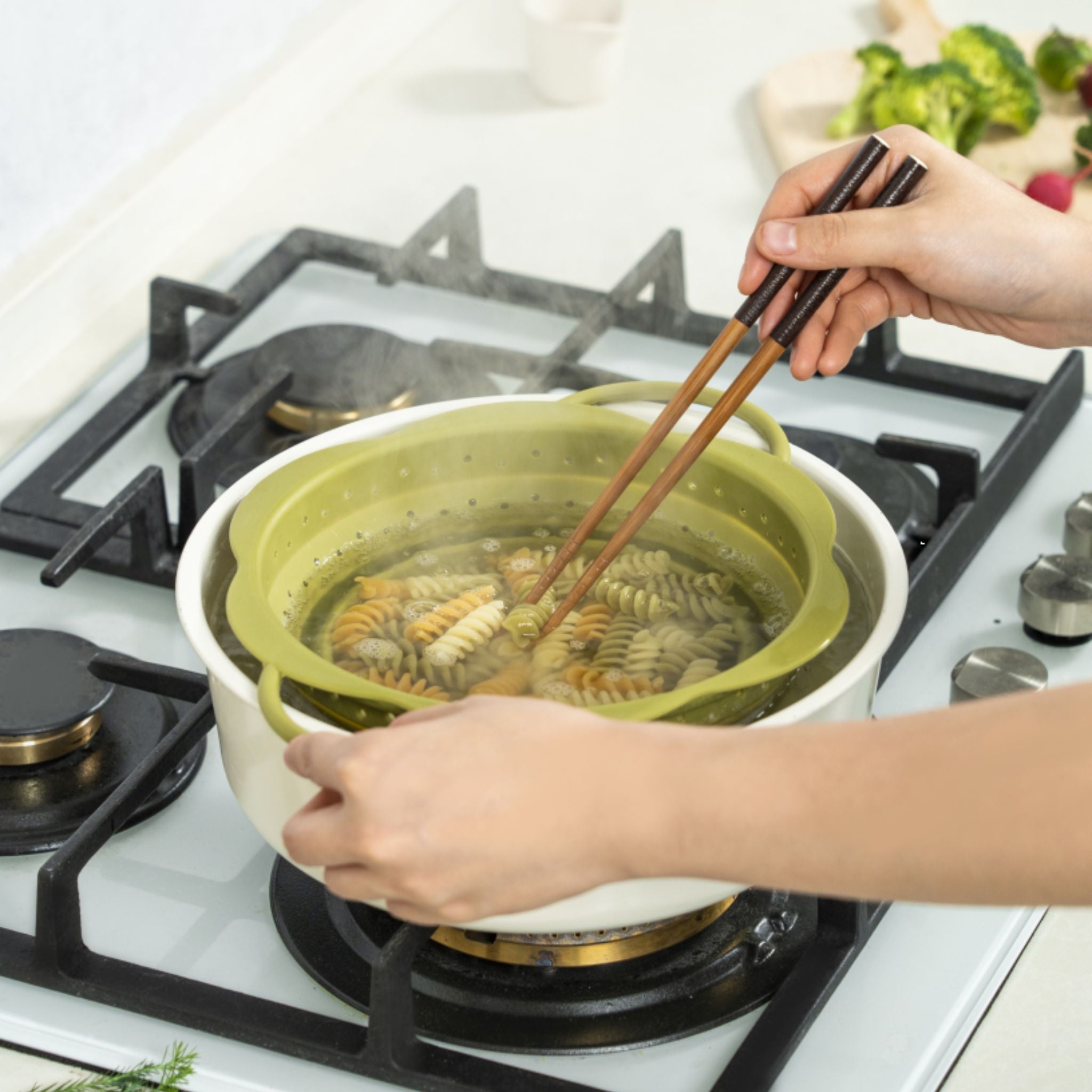 Grünes Faltsieb aus Silikon mit Henkel beim Abseihen von Pasta auf Herd.