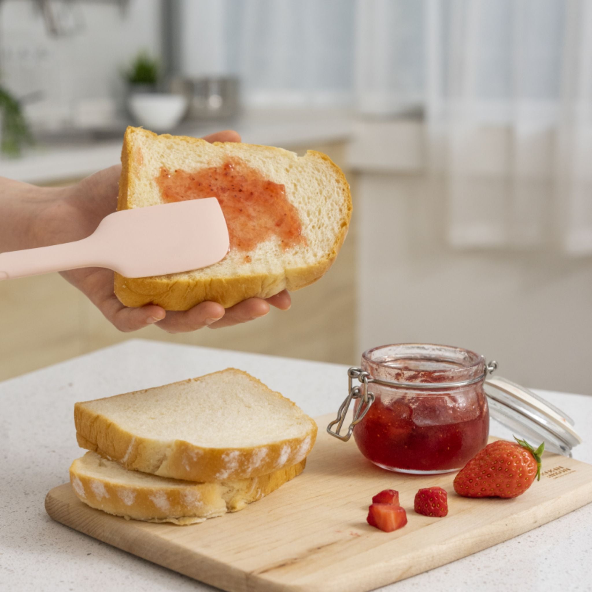Altrosa Teigschaber mit flexiblem Kopf streicht Erdbeermarmelade auf Weißbrotscheibe.