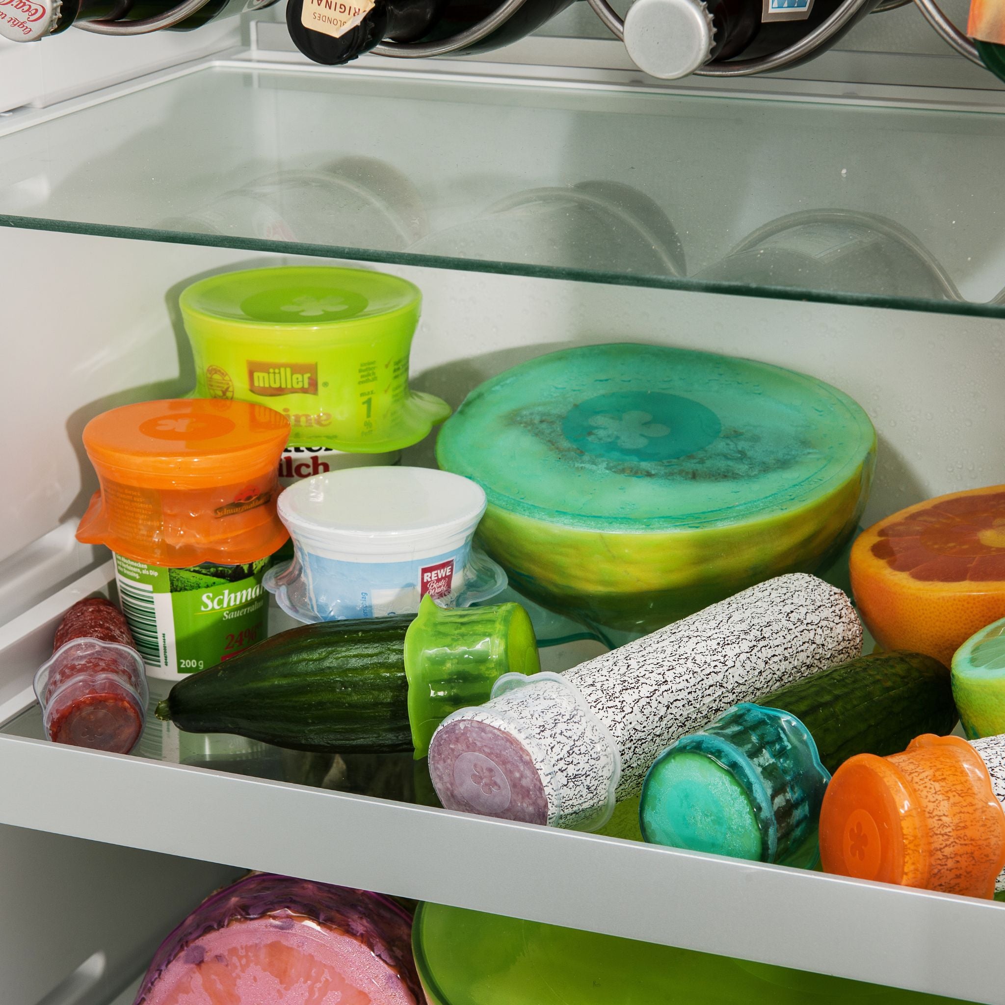 Verschiedene bunte Stretch-Deckel in Orange, Limette, Türkis und Eisblau auf Schüsseln und Lebensmitteln im Kühlschrank.