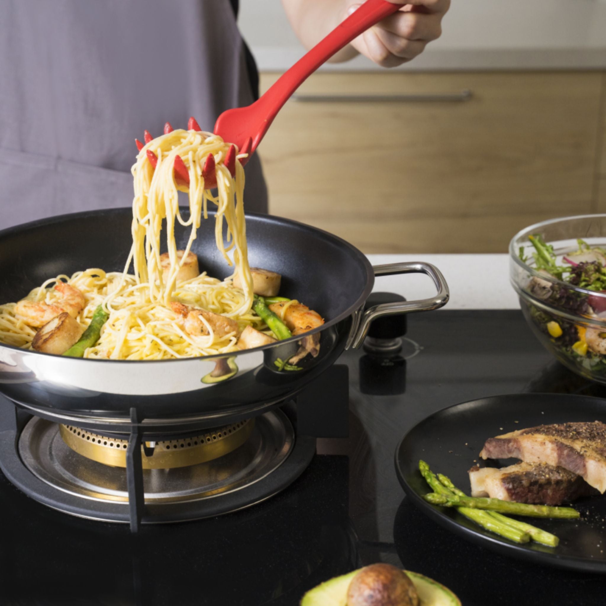 Roter Pastalöffel hebt Spaghetti mit Soße aus schwarzer Pfanne auf Herd.
