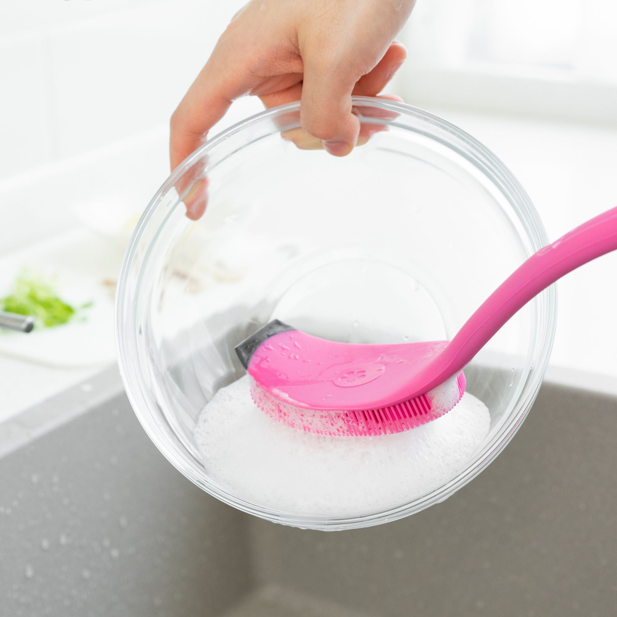 Pinkfarbene Spülbürste mit weichen Silikonborsten reinigt eine Glas-Schüssel über der Spüle.
