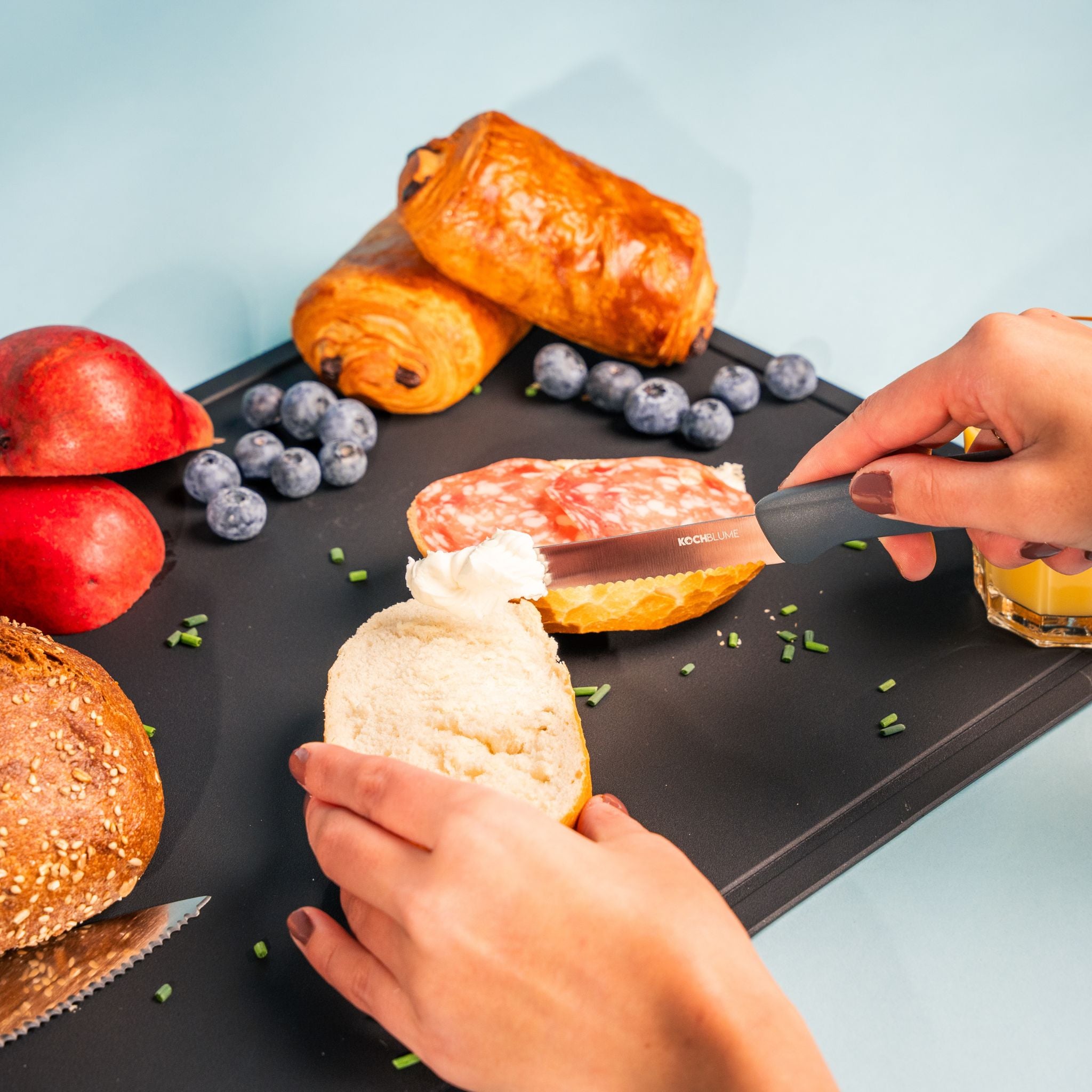 Frühstücksmesser mit schwarzem Griff schneidet Brötchen auf schwarzem Schneidebrett.